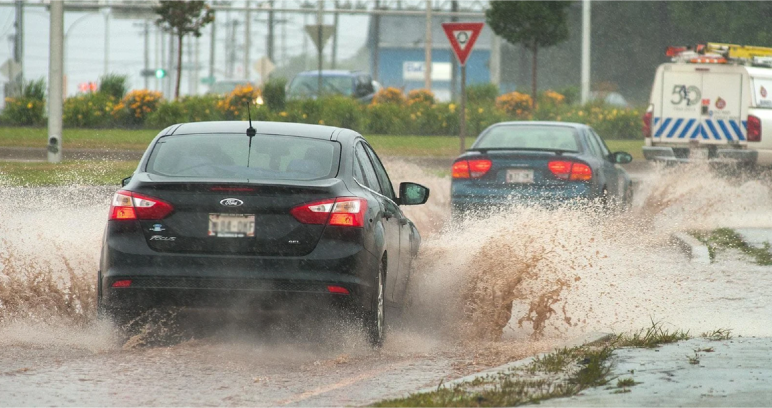 Cars driving through deep, splashing muddy floodwater on a flooded road during heavy rain.