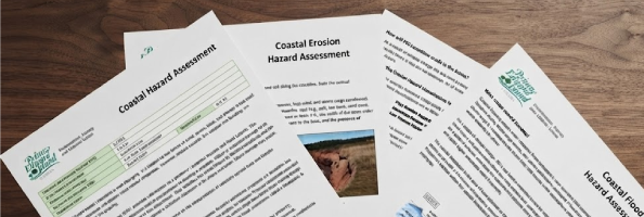Several printed Prince Edward Island documents, including a 'Coastal Hazard Assessment' and 'Coastal Erosion Hazard Assessment,' spread out on a wooden surface.